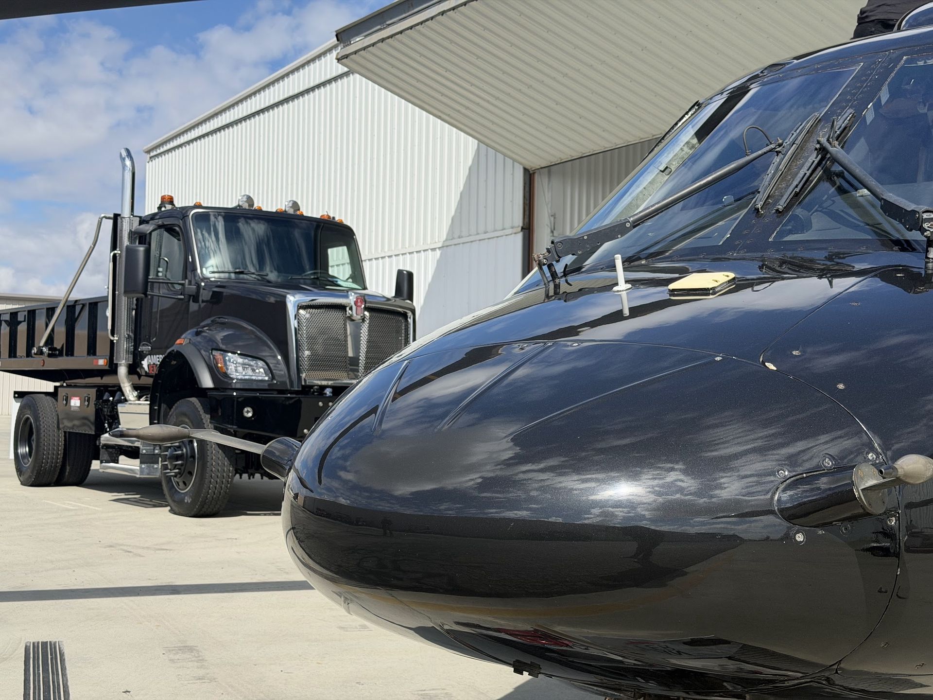 Close‑up view of a glossy black helicopter nose parked outside a hangar, with a black dump truck positioned behind it on a concrete apron | American Jet Center